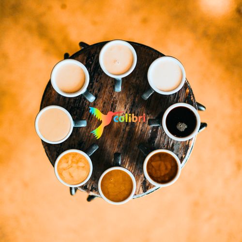 Auf einem runden Holzbrett stehen mit Heißgetränken gefüllt Tassen. In der Mitte steht colibri. Ein bunter Vogel ist abgebildet. Café Colibri - Deutsch sprechen online